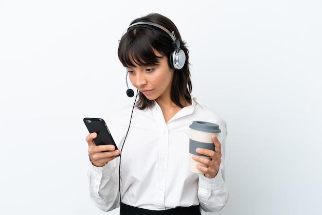 Donna di razza mista di telemarketing che lavora con un auricolare isolato su sfondo bianco che tiene il caffè da portare via e un cellulare