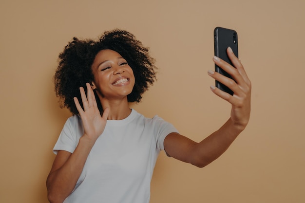 Donna di razza mista felice positiva che saluta con la mano alla telecamera durante la videochiamata su smartphone moderno