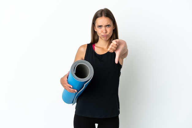 Donna di sport che va alle lezioni di yoga mentre si tiene una stuoia sul muro bianco isolato che mostra il pollice verso il basso con espressione negativa