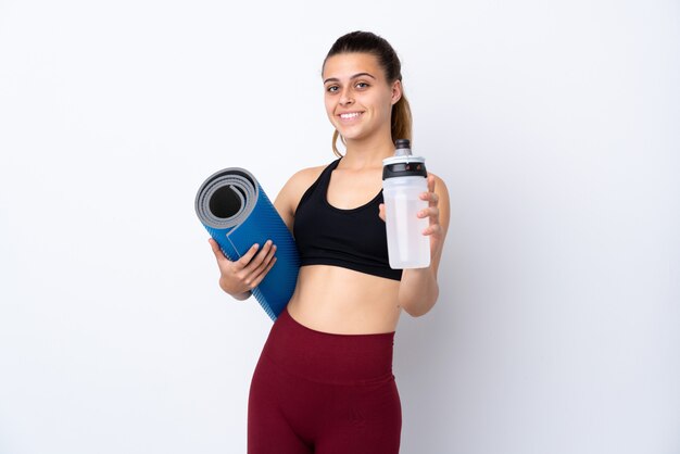 Donna di sport dell'adolescente sopra la parete bianca isolata con la bottiglia di acqua di sport e con una stuoia