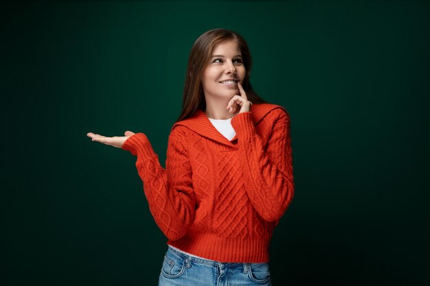 Donna di un anno sognante con i capelli castani in un maglione rosso su uno sfondo verde scuro