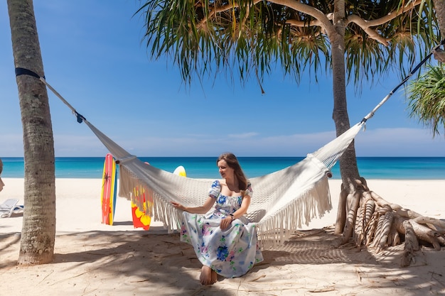 Donna di viaggio che si siede sull'amaca bianca sulla spiaggia sabbiosa con il mare e gli alberi tropicali