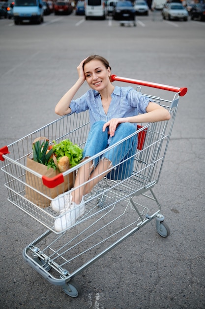 Donna divertente che si siede nel carrello sul parcheggio del supermercato. Cliente felice con acquisti nel centro commerciale, veicoli sullo sfondo