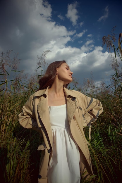 Donna dolce sognante in un impermeabile beige in natura Giovane donna romantica in erba alta spessa nel campo Bellezza naturale godimento della vita