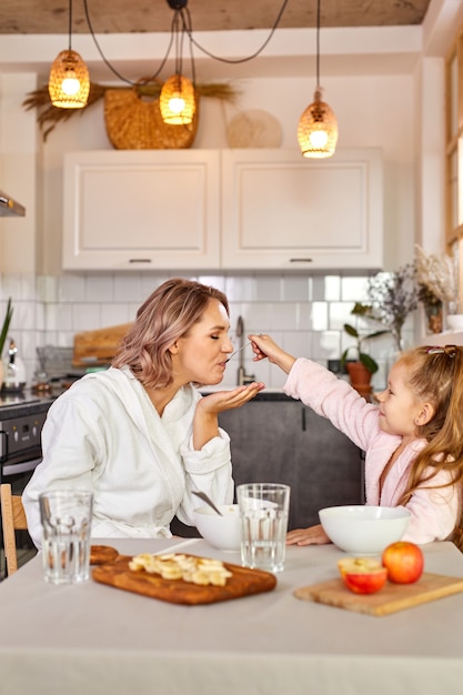 Donna e bambino ragazza fanno colazione in cucina a casa, bella famiglia si siedono insieme mangiando meak, godendo la mattina insieme, divertirsi, sorridere