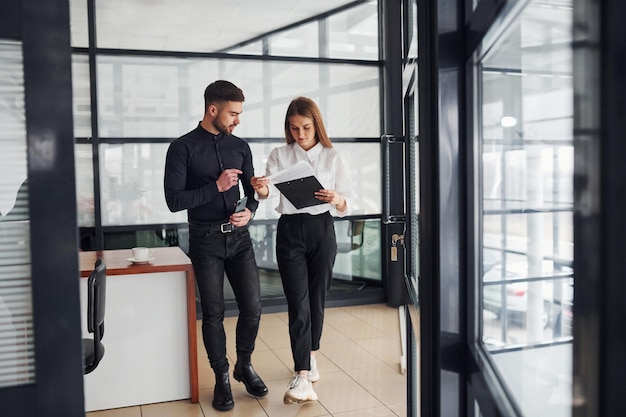 Donna e uomo in abiti formali con documenti che parlano tra loro in ufficio.