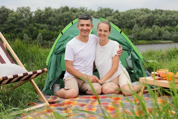 Donna e uomo ottimisti sorridenti che indossano abiti casual seduti alla tenda vicino al fiume che guarda l'obbiettivo facendo un picnic godendosi le giornate primaverili