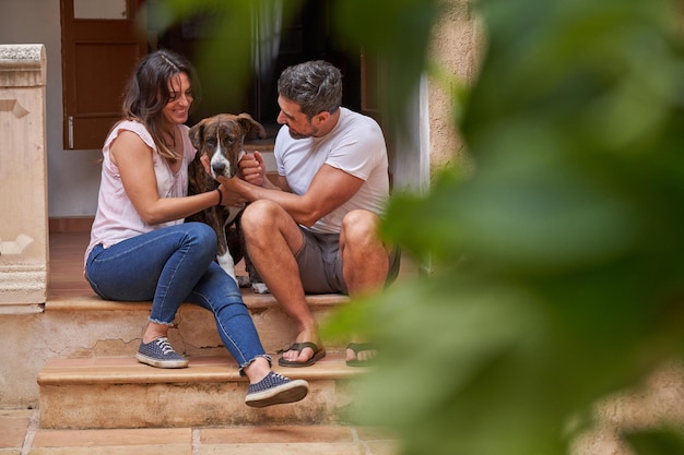 Donna e uomo sorridenti in abiti casual che si divertono seduti sulla veranda e accarezzano insieme un adorabile cane a casa