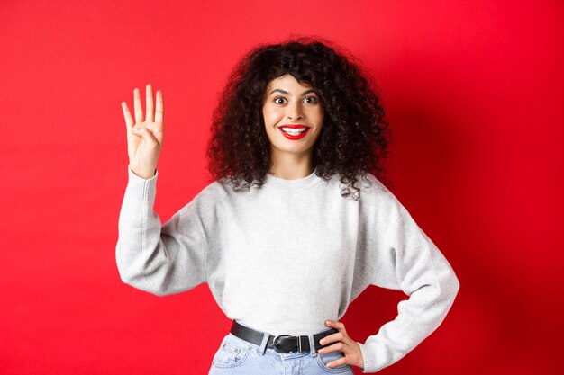 Donna eccitata con capelli ricci che mostra il numero quattro con le dita, facendo ordine, in piedi su sfondo rosso.
