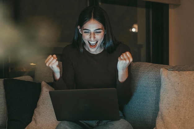 Donna eccitata esultare e festeggiare di fronte a un portatile aperto seduto sul divano a casa vincitore persone femminili ha espressione gioiosa guardando computer display successo e felicità momento interno