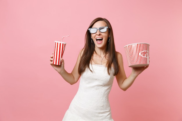 Donna eccitata in abito bianco, occhiali 3d che guardano film, tenendo in mano un secchio di popcorn tazza di plastica di soda o cola