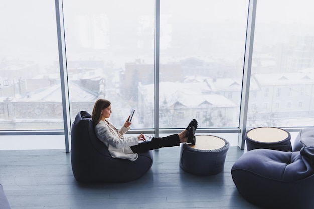 Donna elegante che analizza i grafici sui gadget e si concentra sullo schermo del telefono mentre è seduta vicino alla finestra a tutta parete in un ufficio moderno