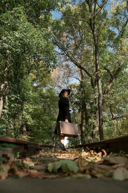 Donna elegante che cammina su rotaie che camminano attraverso la foresta Cappello a tesa larga cappotto nero e valigetta retrò in mano Vista dal basso