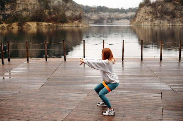 Donna elegante e sportiva con i capelli rossi in abbigliamento sportivo squat con banda elastica fitness su un molo di legno vicino al lago con rocce concetto sportivo stile di vita sano