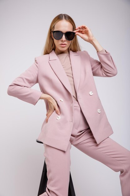 donna elegante in abito rosa pallido piuttosto pastello, giacca, pantaloni, occhiali da sole su sfondo bianco