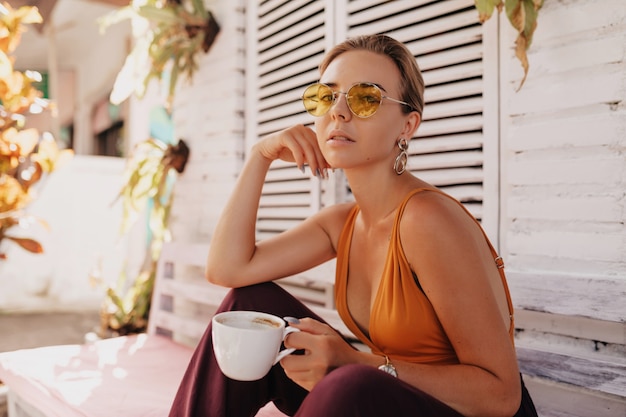 Donna elegante romantica con capelli biondi che indossa occhiali da sole alla moda rotondi e maglietta arancione che si siedono fuori con caffè