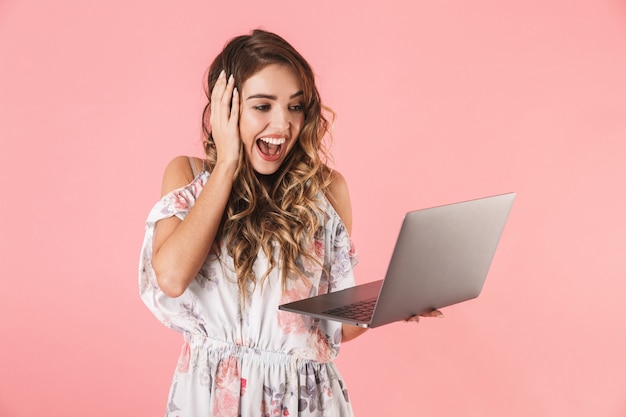 donna emotiva in abito azienda e utilizzando laptop argento, isolato in rosa