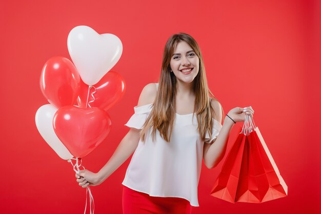 Donna emozionante con i sacchetti della spesa e palloni a forma di cuore isolati sopra la parete rossa