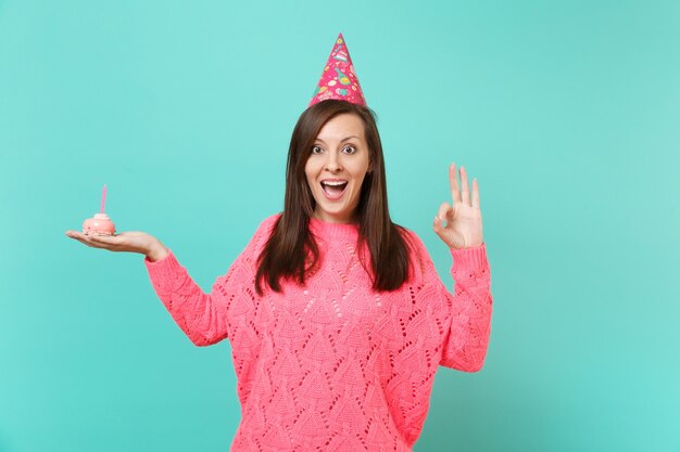 Donna emozionante in maglione rosa lavorato a maglia, cappello di compleanno che tiene la bocca spalancata, mostrando il gesto OK tenere in mano la torta con la candela isolata su sfondo blu. Concetto di stile di vita della gente. Mock up copia spazio.