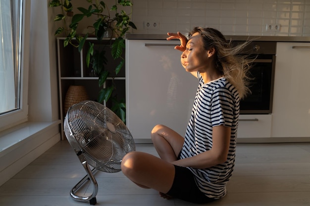 Donna esausta rinfrescante sedersi vicino al grande ventilatore interno che soffia aria fresca di raffreddamento a casa sul pavimento