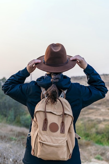Donna escursionista dalla parte posteriore con giacca blu e zaino in spalla tiene il suo cappello hipster mentre guarda l'orizzonte