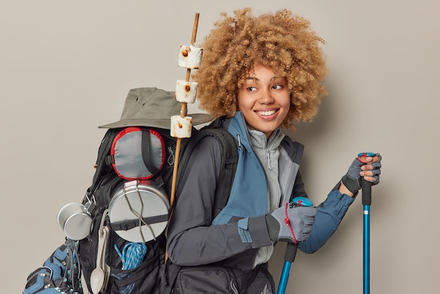 Donna escursionista ha capelli ricci passeggiate con bastoncini da trekking torna dal picnic porta zaino con utensili attrezzature e marshmallow arrosto coinvolto in attività di campeggio isolato su sfondo grigio