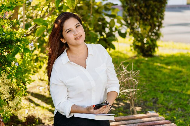 Donna esecutiva che si siede con un libro usando il telefono su una panchina del parco