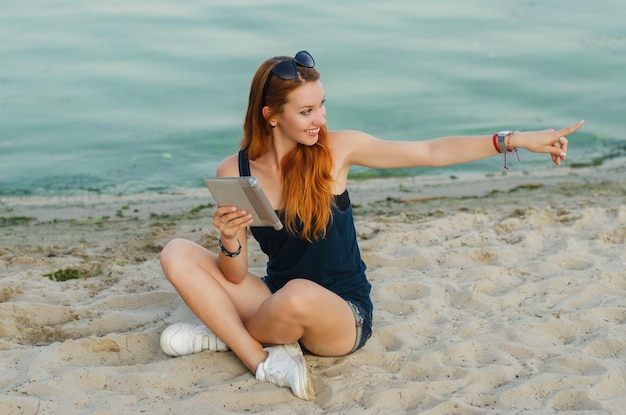 Donna esile sorridente della testarossa che si siede su una spiaggia e che tiene il computer della compressa.