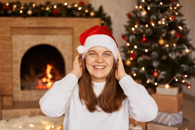 Donna estremamente felice che indossa un maglione bianco e un cappello di babbo natale, in posa vicino al camino e all'albero di natale, guardando la telecamera con emozioni positive, celebrando il Natale.