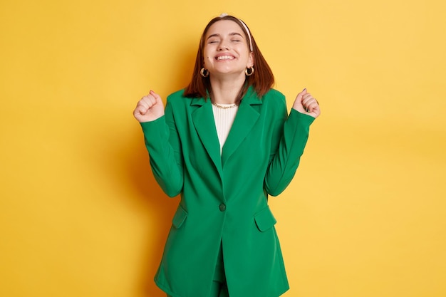 Donna estremamente felice che indossa una giacca verde in piedi isolata su sfondo giallo chiuse gli occhi pugni chiusi celebrando il suo successo