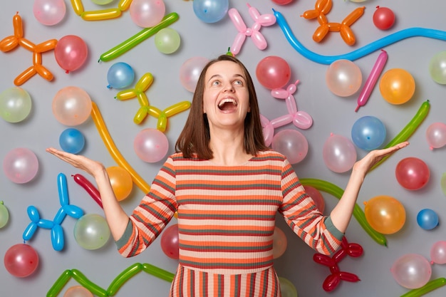 Donna estremamente felice e felicissima che indossa un abito a righe in piedi contro il muro grigio con decorazione di palloncini che guarda in alto celebrando urlando di felicità e gioia