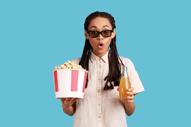 Donna estremamente stupita con i dreadlocks in occhiali 3d imax guardando film film tenere popcorn e bere guardando film incredibili indossando una camicia bianca Colpo in studio al coperto isolato su sfondo blu