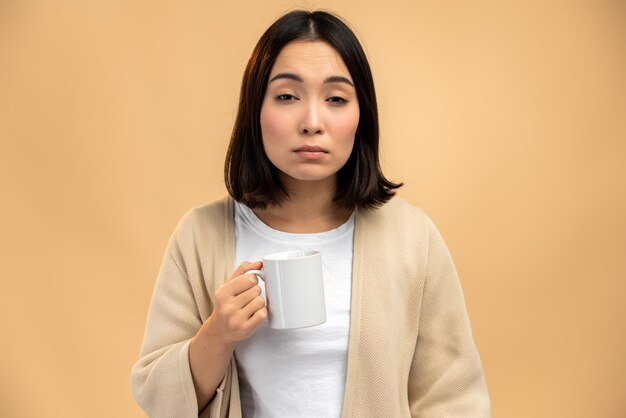 Donna etnica asiatica assonnata con occhi assonnati che tiene la tazza mentre beve caffè al mattino presto su sfondo beige