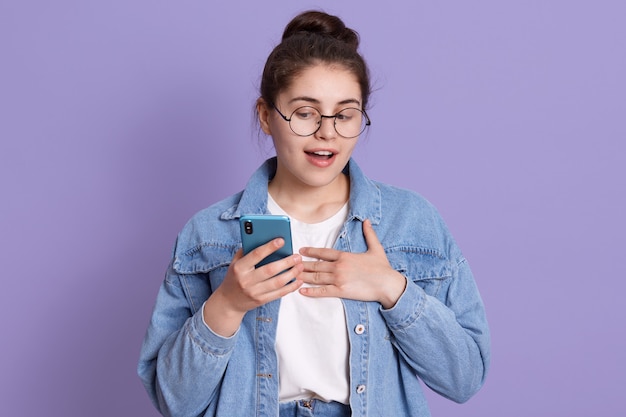 Donna europea emotiva stupita e sorpresa che guarda smart phone con piacevole stupore e gioiosa, indossa giacca di jeans, camicia bianca e occhiali.