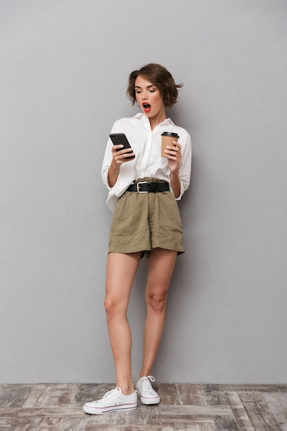 Donna europea emozionante che tiene caffè da asporto e utilizza lo smartphone, isolato sopra il muro grigio