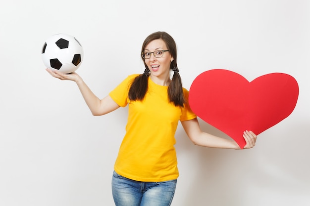 Donna europea sorridente, code di cavallo divertenti, tifoso di calcio o giocatore con gli occhiali, uniforme gialla in possesso di un pallone da calcio classico, cuore rosso isolato su sfondo bianco. Sport, calcio, concetto di stile di vita sano.