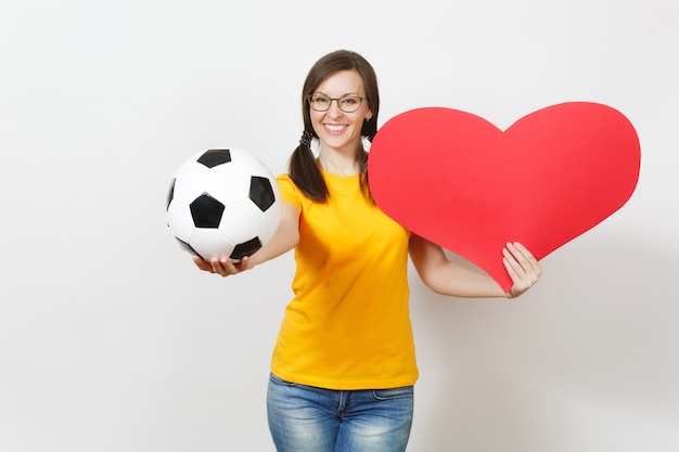 Donna europea sorridente, code di cavallo divertenti, tifoso di calcio o giocatore con gli occhiali, uniforme gialla in possesso di un pallone da calcio classico, cuore rosso isolato su sfondo bianco. Sport, calcio, concetto di stile di vita sano.