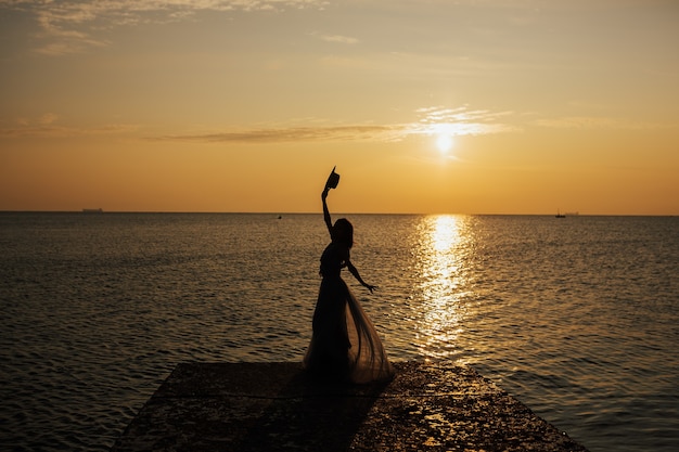 Donna felice al tramonto estivo in mare alza le mani al cielo arancione.