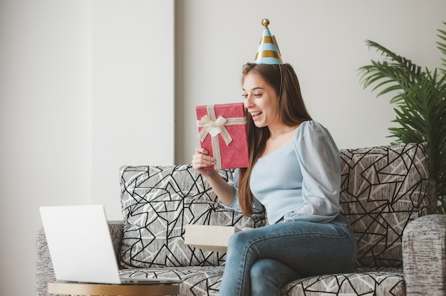 Donna felice che apre la confezione regalo presente e si sente sorpresa durante la nuova normale festa di Natale online a casa sul divano tramite laptop