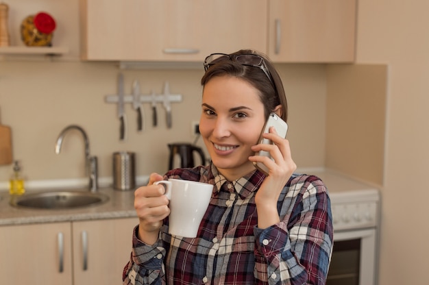 Donna felice che beve il tè in cucina a casa e parlando al telefono