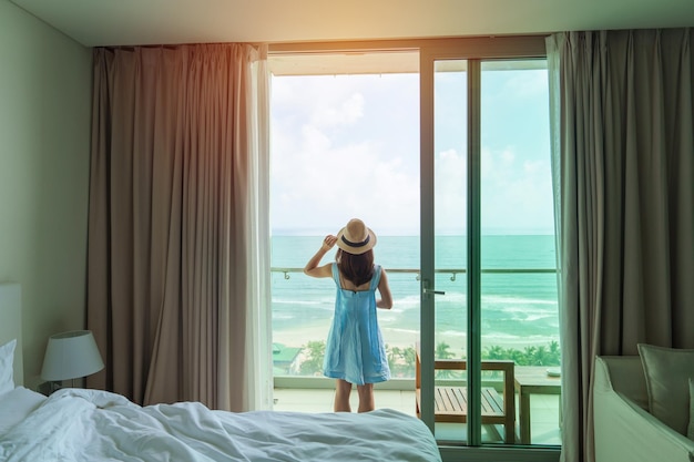 Donna felice che indossa abito blu e cappello che guarda fuori dalla finestra con vista sull'oceano al mattino Rilassati il tempo di vacanza per viaggiare e il concetto di libertà