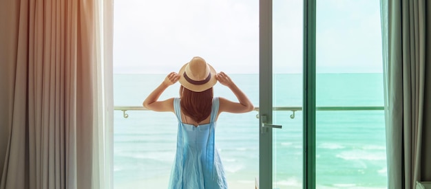 Donna felice che indossa abito blu e cappello che guarda fuori dalla finestra con vista sull'oceano al mattino Rilassati il tempo di vacanza per viaggiare e il concetto di libertà