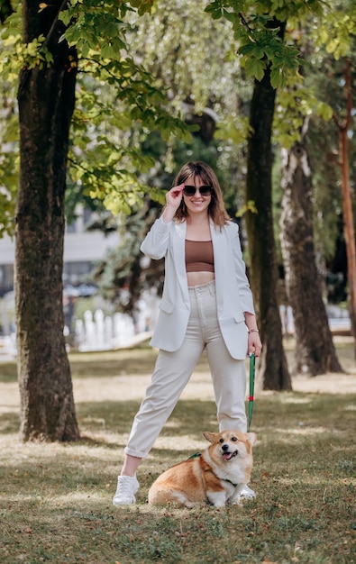 Donna felice che indossa un abito bianco e occhiali da sole che cammina nel parco cittadino con il cane Welsh Corgi Pembroke