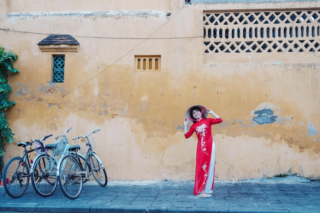 Donna felice che indossa un abito vietnamita Ao Dai e un viaggiatore con cappello che visita la città antica di Hoi An nel punto di riferimento del Vietnam centrale e popolare per le attrazioni turistiche Concetto di viaggio del Vietnam e del sud-est