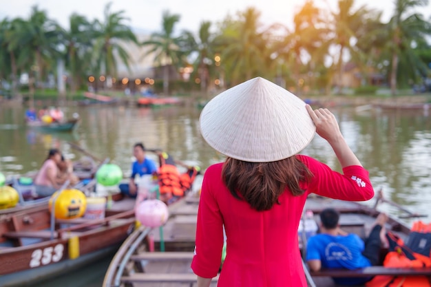 Donna felice che indossa un abito vietnamita Ao Dai, il viaggiatore visita il fiume Thu Bon e il giro turistico in barca a Hoi An, un punto di riferimento della città antica per le attrazioni turistiche, concetto di viaggio in Vietnam e nel sud-est