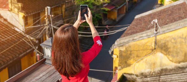 Donna felice che indossa un abito vietnamita Ao Dai viaggiatore che scatta foto con il telefono cellulare sul tetto di Hoi An antica città in Vietnam Vietnam e concetto di viaggio sud-est