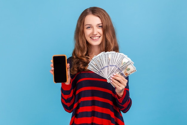 Donna felice che indossa un maglione in stile casual a righe, mostrando fan di banconote in dollari e telefono cellulare con schermo nero vuoto per la pubblicità. Studio indoor girato isolato su sfondo blu.