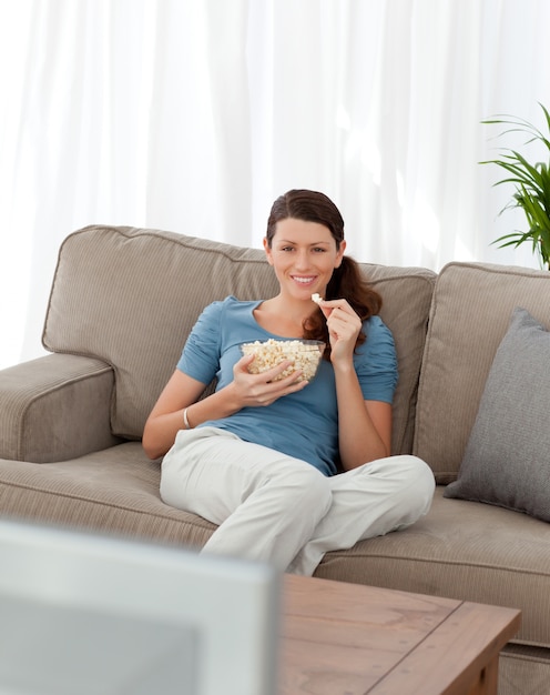 Donna felice che mangia pop corn mentre si guarda la televisione sul divano