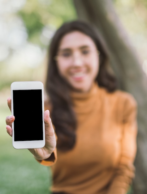 Donna felice che mostra Smart Phone in bianco Schermo sopra il fondo del parco.