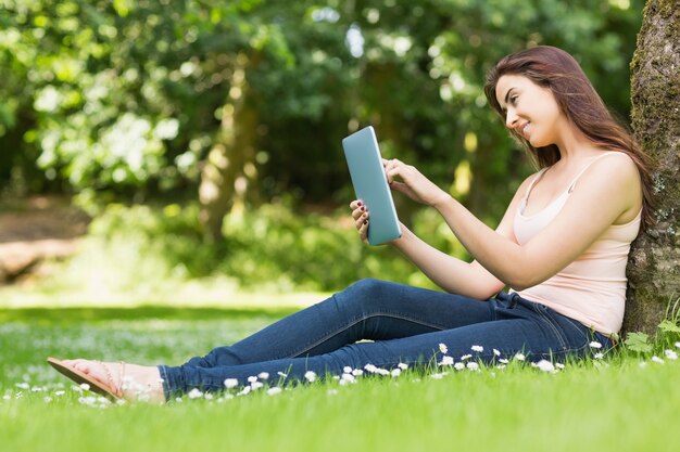 Donna felice che si appoggia contro un albero che lavora con il suo tablet